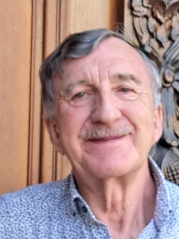 A smiling older man with a mustache and graying hair looks directly at the viewer.