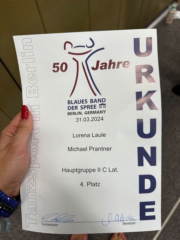 Hand holding a German certificate, celebrating 50 years, with names and "4. Platz" visible.