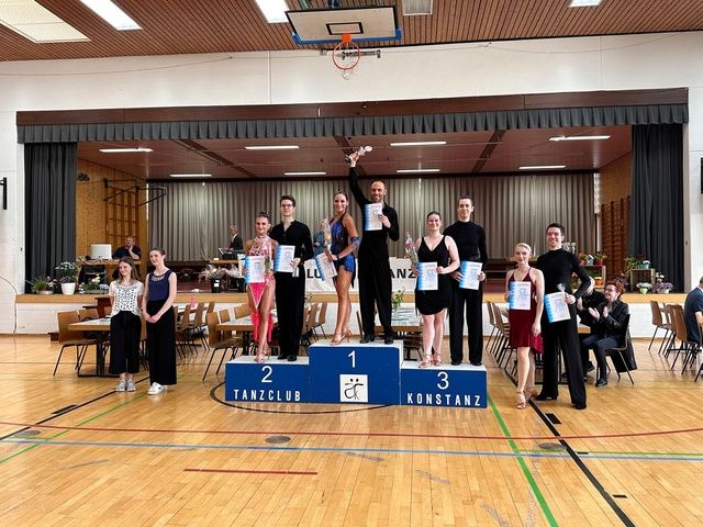 Ballroom dancers in formal wear stand on a podium with certificates after a competition.
