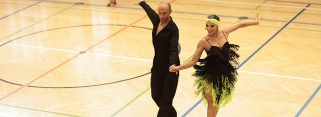 A man and woman in dance attire perform a ballroom dance on a wooden floor.