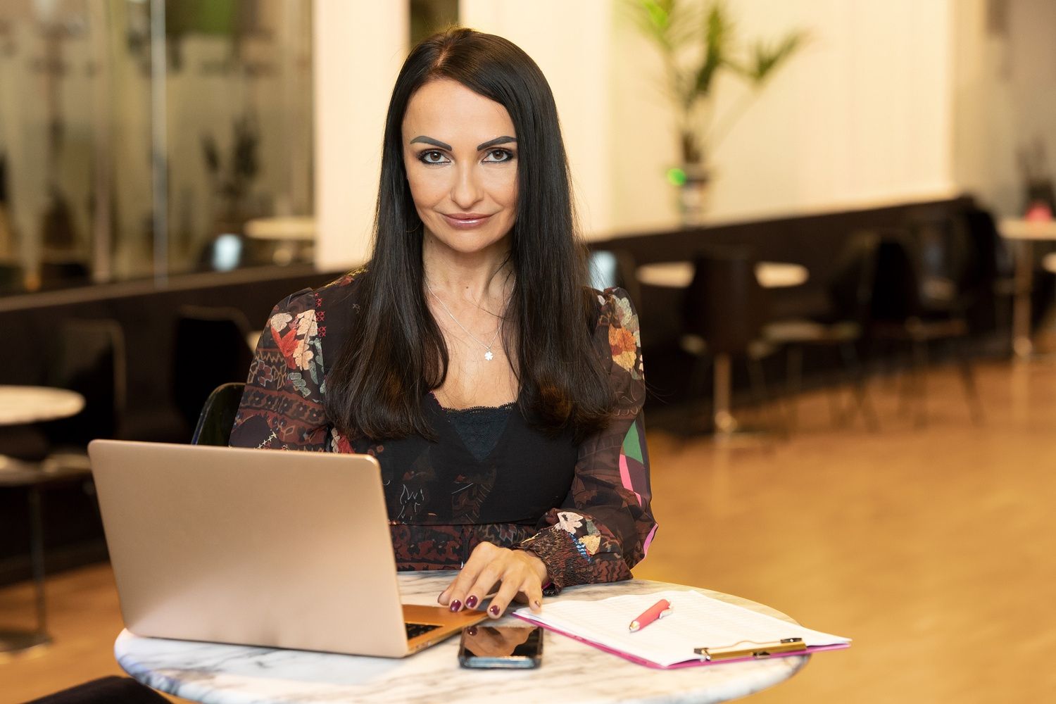 A woman with dark hair sits at a table, working on a silver laptop.