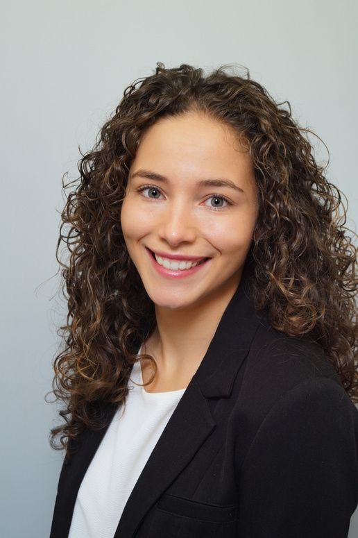 A young woman with long curly brown hair and a black blazer smiles warmly.