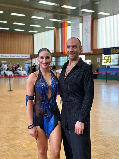 A male and female ballroom dancer pose in their competitive attire inside a hall.