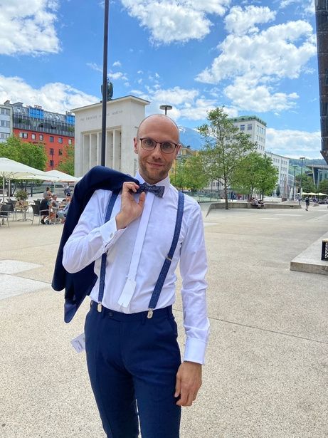 A stylish bald man with glasses, a bow tie, and suspenders poses outdoors.