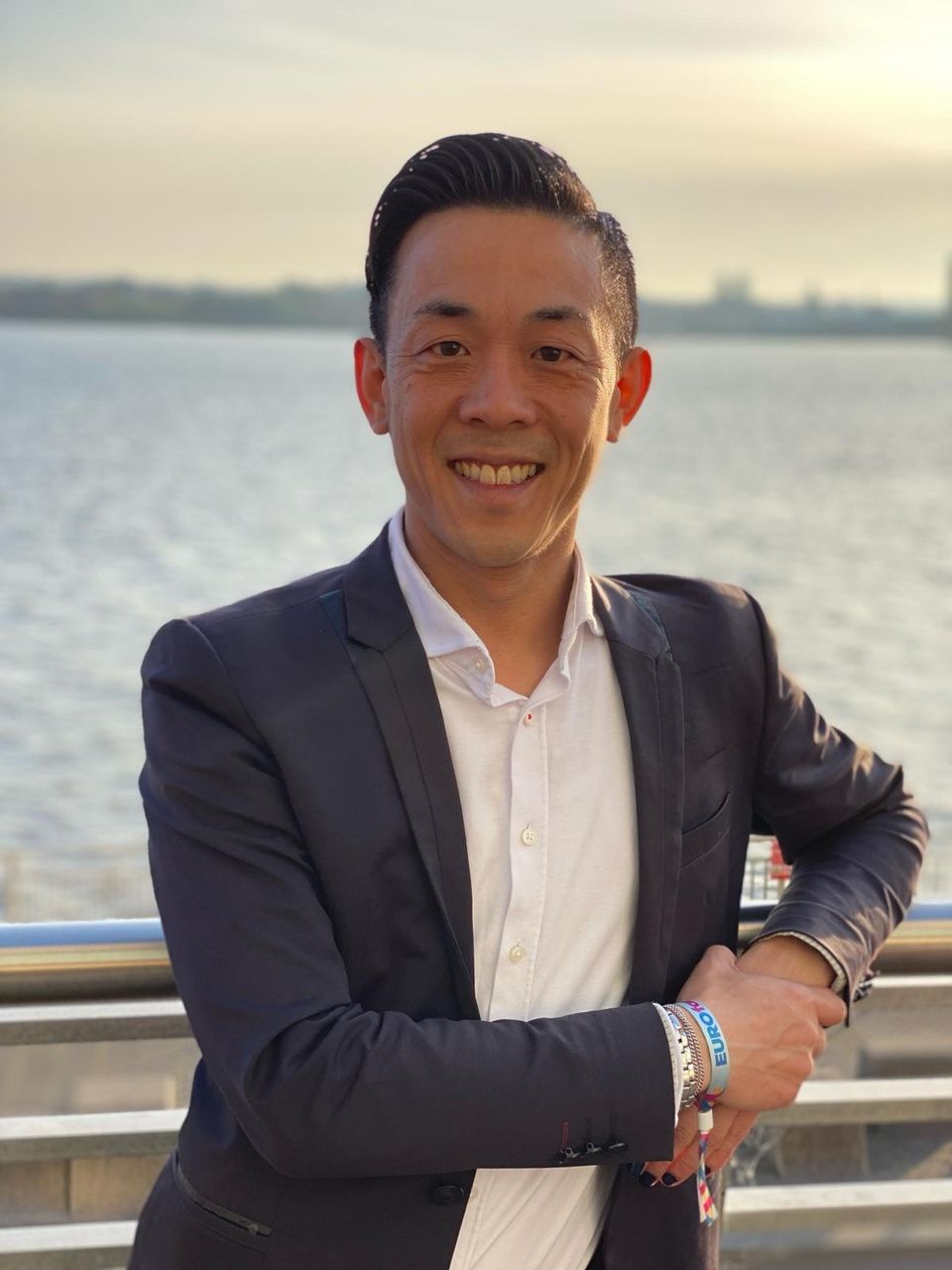 A smiling Asian man in a suit jacket stands outdoors by water.