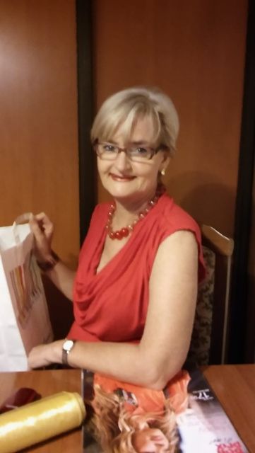 A smiling woman with short blonde hair and glasses holds a white gift bag.